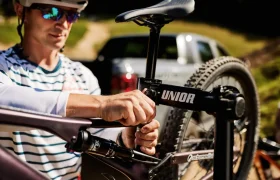 Una persona, con casco y gafas de sol, utiliza herramientas Unior para ajustar el asiento de una bicicleta. Al fondo, un coche y árboles aportan un toque urbano y tranquilo.