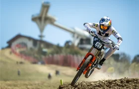 Un ciclista de montaña, vestido con un maillot de competición blanco y casco, y equipado con el TRP EVO PRO Anniversary Edition, desciende por una pista de tierra, con un gran salto de esquí y un edificio visibles al fondo.