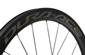 Primer plano de una llanta de bicicleta de carbono negro con la marca «DURA-ACE» en letras grandes, en la que se aprecian varios radios plateados. Este modelo Dura-Ace de Shimano, el WH-R9370, destaca por la precisión y la ligereza características de las ruedas de carbono.