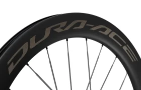 Primer plano de una rueda de bicicleta con llanta negra y la marca «DURA-ACE» en letras grandes, con radios plateados sobre un fondo blanco. Este modelo Shimano Dura-Ace WH-R9370 destaca por sus ruedas de carbono de alto rendimiento, diseñadas para ofrecer velocidad y precisión.
