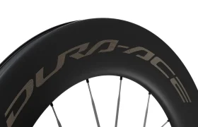 Primer plano de una rueda de bicicleta de carbono negro con radios plateados, en la que destaca la distintiva marca «DURA-ACE» en la llanta, escrita con letras grandes y estilizadas. Esta rueda Shimano Dura-Ace WH-R9370 destaca por su avanzado diseño de radios de carbono, que mejora el rendimiento y la durabilidad.