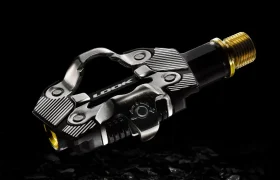 Un pedal de bicicleta negro y plateado con un eje dorado del ecosistema de pedales LOOK se muestra sobre un fondo oscuro y texturizado.