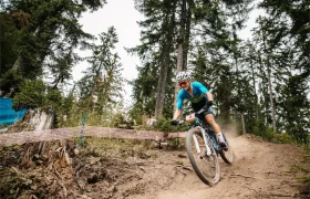 Jenny Rissveds, con maillot y casco azules, conduce una bicicleta de montaña por un sendero de tierra en una zona boscosa.
