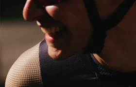 Primer plano de una persona que lleva un casco de bicicleta, gafas de sol y una camiseta sin mangas de malla «Castelli Transition», sonriendo y mirando ligeramente hacia un lado, bajo luz natural.
