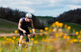 Un ciclista que lleva un casco Castelli Transition y unas gafas de sol recorre en bicicleta un paisaje cubierto de hierba con flores silvestres amarillas en primer plano, vestido con un maillot Corretto LS de manga larga.