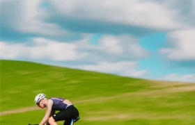 Un ciclista con equipación de competición, que lleva un maillot Corretto LS, pedalea a gran velocidad en una bicicleta de carretera por un paisaje cubierto de hierba, bajo un cielo azul con nubes dispersas.