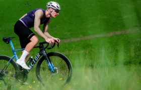 Un ciclista con equipación de competición —que luce un maillot Corretto LS y una camiseta de manga larga Castelli Transition—, pedalea cuesta arriba en una bicicleta de carretera azul por un paisaje cubierto de hierba, con un casco blanco y gafas de sol.