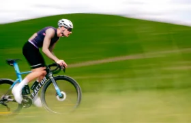 Un ciclista, con casco y gafas de sol, pasa a toda velocidad en su bicicleta de carretera junto a una colina cubierta de hierba, de un verde difuminado, bajo un cielo nublado, luciendo el elegante maillot Corretto LS de la colección «manga larga» de Castelli.