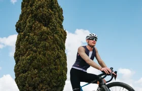 Un ciclista con gafas de sol y casco se detiene en su bicicleta de carretera frente a un ciprés alto, bajo un cielo azul con nubes dispersas, luciendo el maillot Corretto LS para disfrutar de una comodidad óptima en este recorrido panorámico.