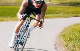 Un ciclista que lleva casco y gafas de sol, vestido con un maillot Corretto LS, se inclina al tomar una curva en una carretera rural rodeada de hierba y campos abiertos, bajo un cielo parcialmente nublado.