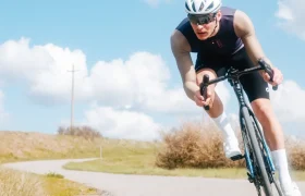 Un ciclista, con casco y gafas de sol, circula en bicicleta por una carretera sinuosa al aire libre, bajo un cielo azul con algunas nubes dispersas, luciendo el maillot de manga larga Corretto LS para disfrutar de una comodidad óptima.