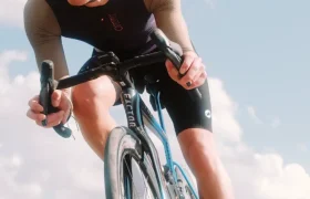 Un ciclista que lleva casco, gafas de sol y ropa de ciclismo, incluido el maillot Corretto LS, circula en bicicleta de carretera por un camino asfaltado bajo un cielo parcialmente nublado.