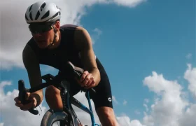 Un ciclista que lleva un casco blanco, gafas de sol y un maillot Corretto LS pedalea en una bicicleta de carretera al aire libre bajo un cielo azul parcialmente nublado.