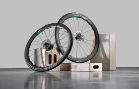 Dos ruedas de bicicleta de carbono Zipp 2026 con frenos de disco se muestran apoyadas sobre cajas de madera sobre una superficie lisa y reflectante, listas para rodar más rápido contra el fondo gris liso.