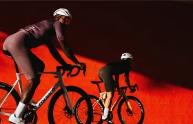 Dos ciclistas, con casco y equipación de ciclismo, pedalean en sus bicicletas de carretera Zipp 404 S junto a una pared de un rojo intenso, iluminados por un espectacular rayo de sol que evoca tanto la velocidad como el estilo.