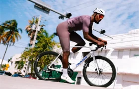 Un ciclista, que lleva casco y gafas de sol, pedalea en una bicicleta de carretera Colnago blanca con ruedas Zipp 404 S, disfrutando de la velocidad por una calle urbana soleada, flanqueada por palmeras y edificios.