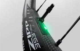 Primer plano de una rueda de bicicleta de carbono Zipp 2026 con un dispositivo sensor iluminado en verde adjunto, que muestra conectividad inteligente y dos radios visibles, diseñados para ayudarte a rodar más rápido.