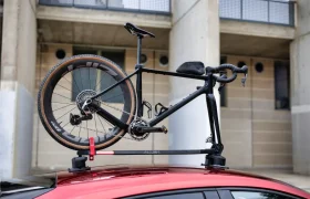 Una bicicleta de carretera negra montada boca abajo en un portabicicletas Allen Smart Suction Bike Rack sobre un coche rojo, con un edificio de hormigón al fondo.