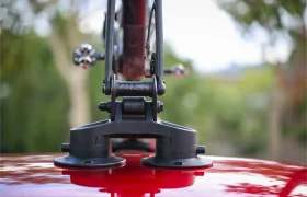 Primer plano de un portabicicletas Allen Smart Suction montado en el techo de un coche rojo, con la marca "Allen" visible. Los árboles aparecen difuminados en el fondo, resaltando el elegante diseño de este portabicicletas Allen Smart Suction.