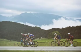 Cuatro ciclistas recorren un camino rural bordeado de campos verdes y colinas boscosas, con una montaña y un cielo nublado al fondo: un escenario ideal para una aventura ciclista de Alpinum TotalEnergies.