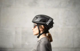 Vista de perfil de una persona que lleva un casco de ciclista POC negro casco ligero, gafas de sol y una camiseta gris, de pie delante de un muro de piedra gris claro.