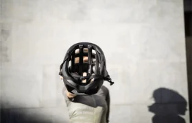 Una persona sostiene un POC Cytal Lite, un elegante casco ventilado, hacia la cámara, oscureciendo su rostro contra una pared gris lisa con una sombra visible a la derecha.