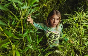 Una persona se encuentra entre densas plantas verdes de bambú, agarrada a una rama y mirando hacia la cámara con expresión neutra, luciendo un maillot ASSOS del equipo EF Pro Cycling, un sutil guiño a las aspiraciones del Giro 2026.