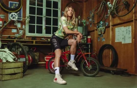 Una mujer vestida con la equipación de EF Pro Cycling se sienta en una pequeña bicicleta BMX roja dentro de un taller lleno de bicicletas, herramientas, neumáticos y bocetos en las paredes de madera, lista para abrazar el espíritu del Giro 2026.