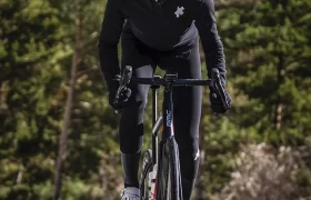 Un ciclista con una chaqueta negra EQUIPE RS Johdah Ultraz ASSOS y casco blanco conduce una bicicleta de carretera por una carretera asfaltada, rodeado de árboles y montañas.