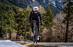 Ciclista con casco y ropa deportiva oscura, incluida la chaqueta extrema EQUIPE RS Johdah Ultraz S11, monta en bicicleta cuesta arriba por una carretera asfaltada, rodeado de árboles y montañas lejanas.