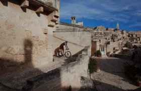 Un ciclista que lleva ASSOS MILLE GT S11 disfruta del confort inteligente mientras recorre un estrecho sendero de piedra en una antigua ciudad en lo alto de una colina con edificios de color canela bajo un cielo azul.