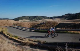Dos ciclistas recorren una carretera asfaltada a través de un paisaje montañoso y seco bajo un cielo azul despejado, mostrando la evolución real y el confort inteligente de la equipación ASSOS MILLE GT S11.