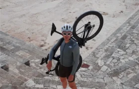 Un ciclista ataviado con una equipación ASSOS MILLE GT S11, casco y gafas de sol, de pie sobre unos escalones de piedra y con una bicicleta al hombro, en un escenario de piedra vacío y al aire libre, muestra a la perfección la evolución real y el confort inteligente.