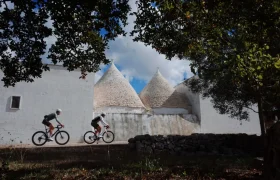 Dos ciclistas con equipamiento ASSOS MILLE GT S11 pasan por delante de tradicionales casas trulli blancas con tejados cónicos, enmarcadas por árboles y un muro de piedra bajo un cielo parcialmente nublado, disfrutando de confort inteligente en su viaje.