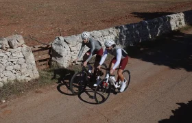 Dos ciclistas con equipamiento ASSOS MILLE GT S11 encarnan la evolución del confort inteligente mientras recorren una carretera rural bordeada por un muro de piedra y campos secos y marrones.
