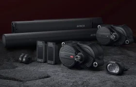 Sobre una superficie de textura oscura y fondo granate se disponen diversos dispositivos y componentes electrónicos de color negro, entre ellos tubos cilíndricos y unidades de control Avinox M2.