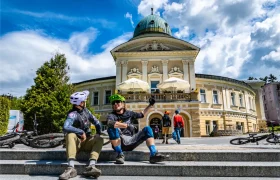 Dos ciclistas con casco y equipación se sientan en unos escalones de piedra con sus bicicletas, charlando frente a un histórico edificio amarillo con una cúpula verde: una escena acogedora en el corazón de Bike Region Glacensis, rodeados de sombrillas al aire libre.