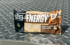 SiS Energy Oat Bar, una barrita energética de chocolate blanco & macadamia con 40g de carbohidratos, reposa sobre una superficie metálica texturizada bajo la luz del sol.