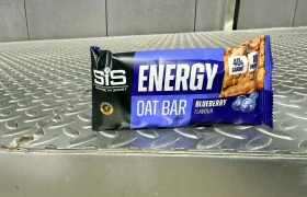 A barrita energética SiS de avena sabor arándano descansa sobre una superficie metálica, mostrando en su etiqueta el aporte de 40g de carbohidratos resaltado en el envase.