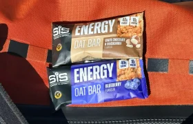 Dos barritas energéticas SiS Energy Oat Bars, una de chocolate blanco y macadamia y otra con sabor a arándanos, se asientan sobre una superficie naranja con una correa negra y una tira reflectante, ideal para un sabroso chute de carbohidratos.