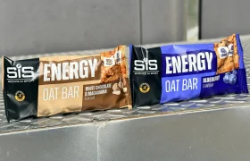 Two SiS Barritas energéticas de avena, una de proteína sabor chocolate blanco y macadamia y otra de arándanos, se exhiben sobre una superficie metálica.