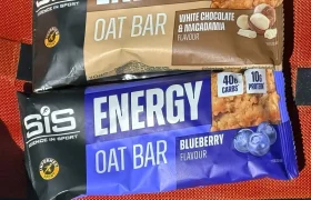 Two SiS barritas energéticas de avena, una de chocolate blanco y macadamia y otra de arándanos, ricas en carbohidratos, se encuentran sobre una tela naranja.