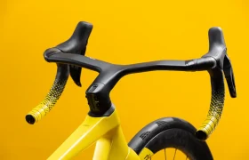 Primer plano del manillar y la rueda delantera de una bicicleta de carretera amarilla sobre un fondo amarillo.