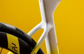 Primer plano de un cuadro y una rueda trasera de una bicicleta de carretera de colores blanco, amarillo y negro sobre un fondo amarillo liso.