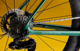 Primer plano del cambio trasero, los piñones y la cadena de una bicicleta sobre un fondo amarillo, con la inscripción «Reparto Corse» impresa en el cuadro verde.