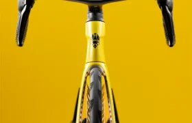En la fotografía se ve una bicicleta de carreras amarilla tomada de frente sobre un fondo amarillo a juego, en la que se aprecian el manillar, la rueda delantera y parte del cuadro.
