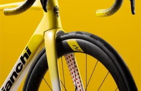 Un primer plano de la parte delantera de una bicicleta de carretera Bianchi amarilla y blanca, en el que se aprecian el manillar, la horquilla con lunares rojos, la rueda y el freno de disco, sobre un fondo amarillo.