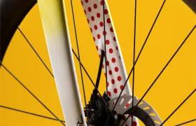 Primer plano de la rueda delantera de una bicicleta con freno de disco y horquilla amarilla, con lunares rojos en la parte interior de la horquilla, sobre un fondo amarillo.