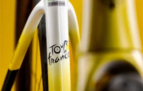 Primer plano de un cuadro de bicicleta amarillo y blanco con el logotipo del «Tour de Francia» y una pegatina de la UCI, sobre un fondo amarillo.