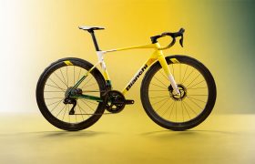 Una bicicleta de carretera con el cuadro en amarillo, blanco y verde, el manillar negro y ruedas de perfil alto, expuesta sobre un fondo con un degradado de amarillo y verde.